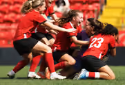 Four female soccer players in red jerseys and black shorts on green field, celebrating while kneeling and embracing each other.