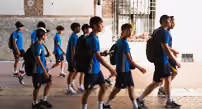 Group of boys wearing blue sports uniforms walking indoors in a line, carrying sports bags.