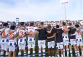 Group of rugby players standing side by side with arms around each other's shoulders facing a large crowd in a stadium.