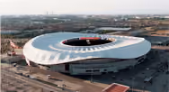 Aerial view of a large, modern, circular stadium with a white roof and an open center, surrounded by empty land and distant buildings.