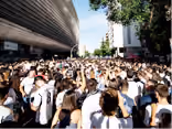 Crowd of people walking through a narrow outdoor space between large buildings under a sunny sky with some trees visible.