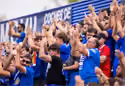 Crowd of sports fans wearing blue shirts cheering with raised hands in a stadium.