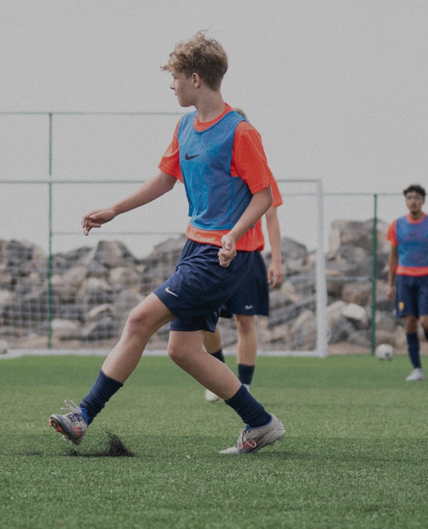 A young male soccer player wearing an orange shirt and blue training bib kicks the ball on a green soccer field with goalposts and rocks in the background, while two other players in similar uniforms stand behind him.