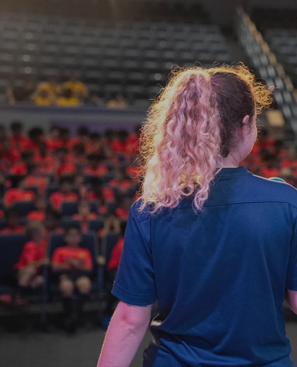 Person with curly, two-toned hair tied in a ponytail, wearing a blue shirt, facing away from the camera in an auditorium with people in red shirts seated in the background.