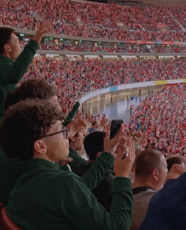 Crowd of spectators enthusiastically cheering and raising hands in a large stadium filled with people, viewed from a close side angle.