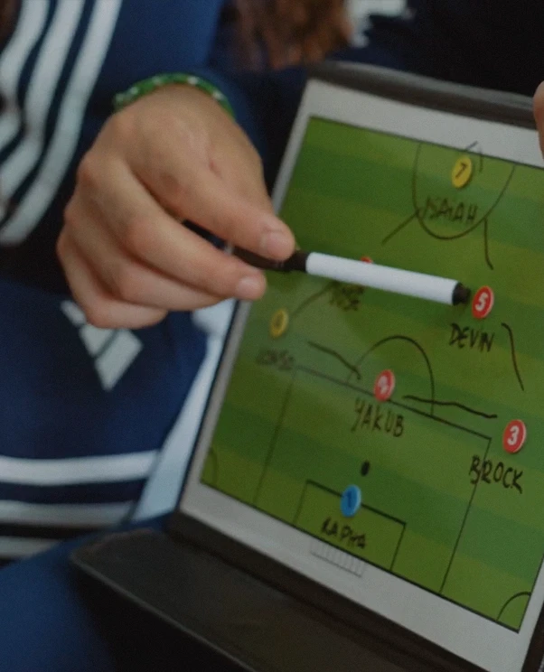 Hand holding a whiteboard marker pointing at a tablet screen showing a soccer field diagram with player names and numbers like Yakub, Devin, Brock, Noah, and La Pita.