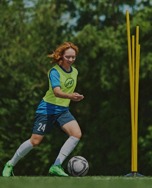 A female soccer player with red hair wearing a yellow training bib, blue shirt, and shorts numbered 24 is dribbling a soccer ball on grass near yellow training poles.