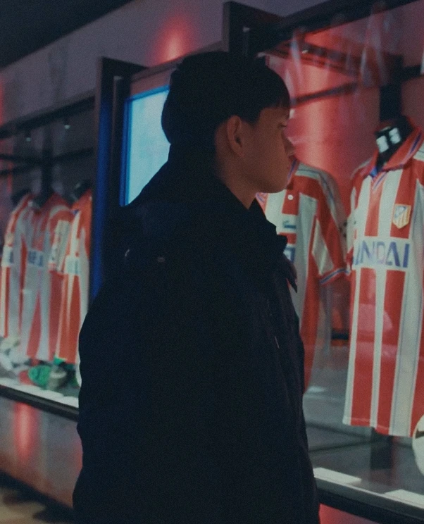Person standing indoors, looking at a display of soccer jerseys with red and white vertical stripes in a glass case.
