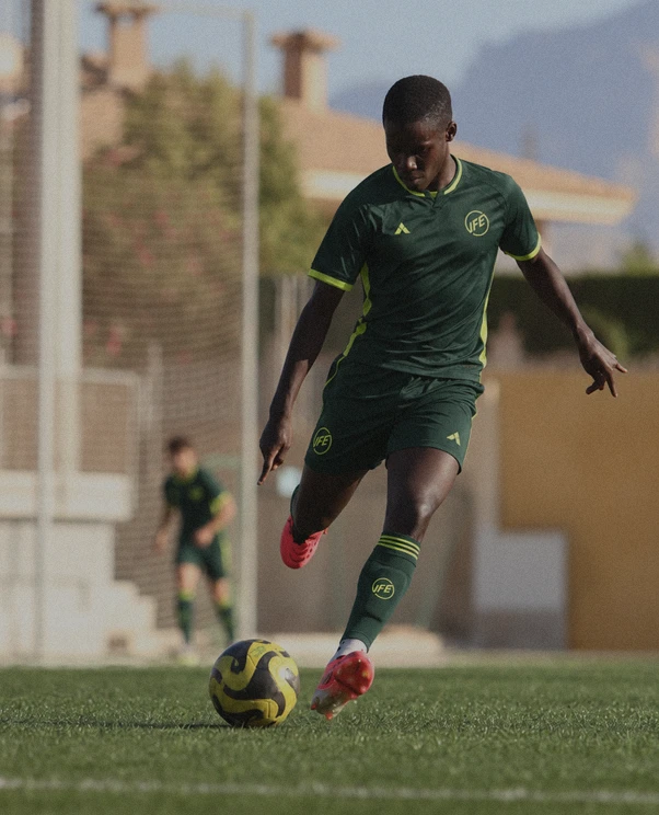 Young soccer player in a green uniform with yellow trim kicking a soccer ball on a grassy field, with another player and buildings blurred in the background.