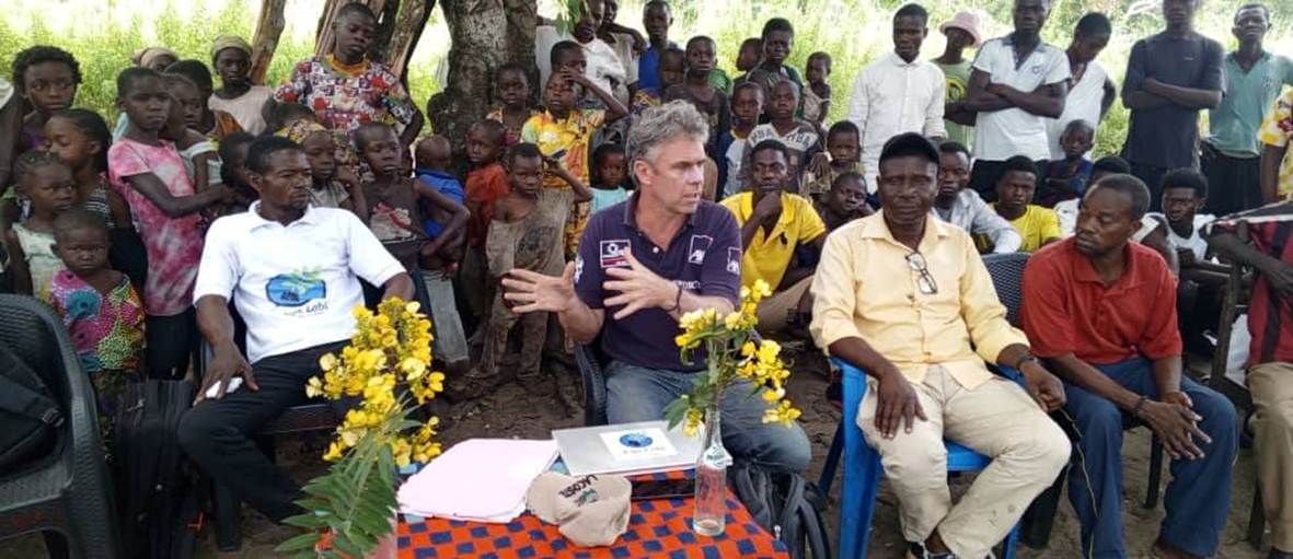 Réunion en plein air : Jurgen Heytens, au centre, assis à une table avec les représentants du village, devant de nombreux habitants rassemblés.