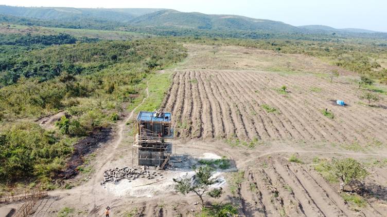 Vue aérienne d’une tour d’eau au bord d’un grand terrain labouré, entre forêt et collines.