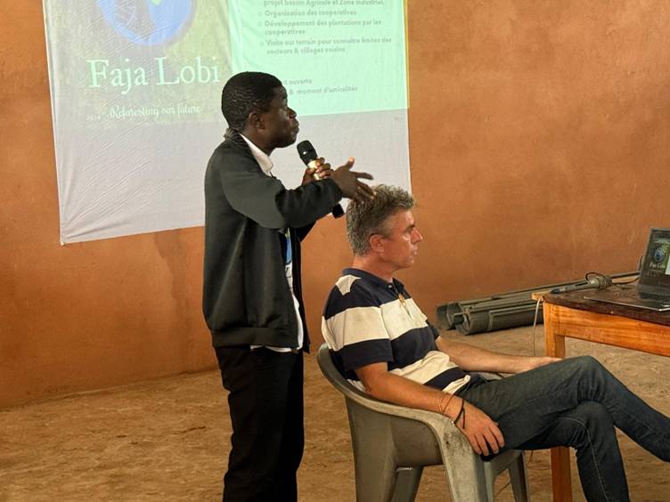 Un intervenant parle au micro devant une projection Faja Lobi, à côté d’un membre de l’équipe assis.