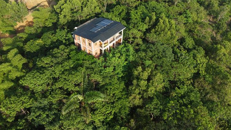Vue aérienne d’une maison au toit équipé de panneaux solaires, entourée d’une forêt dense.