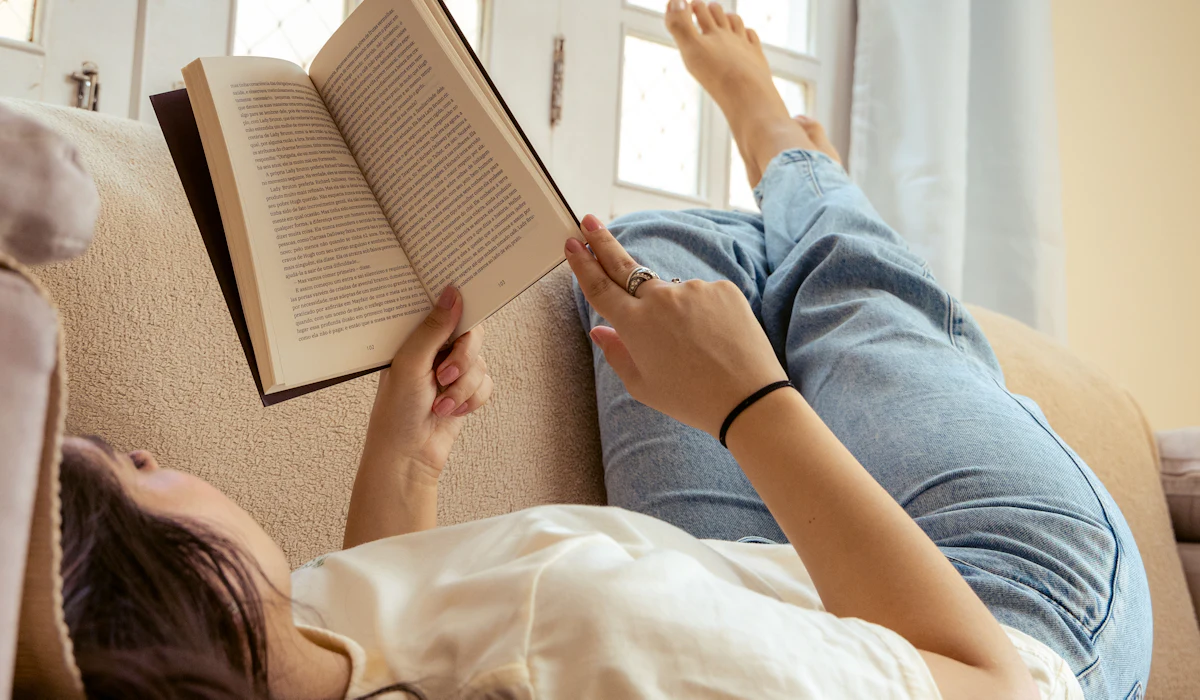 Personne allongée sur un canapé, lisant un livre près d’une fenêtre