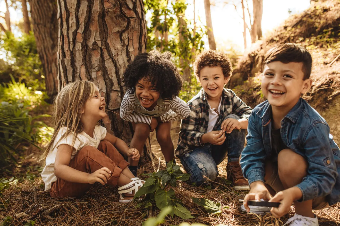 Four laughing children playing together on the forest floor beside a tree