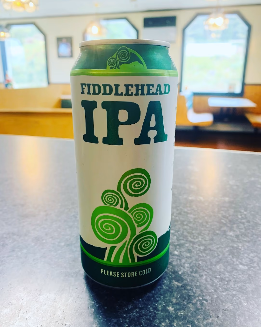 Can of Fiddlehead IPA beer on a table with a green and white label featuring stylized fiddlehead fern illustrations and text saying "Please Store Cold."