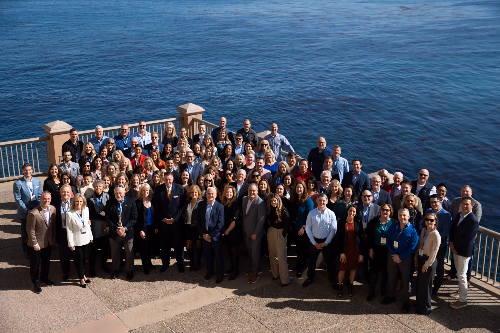 Large group of about 80 people standing on a terrace with a stone floor and metal railing, with ocean water in the background, posing for a group photo under sunlight.