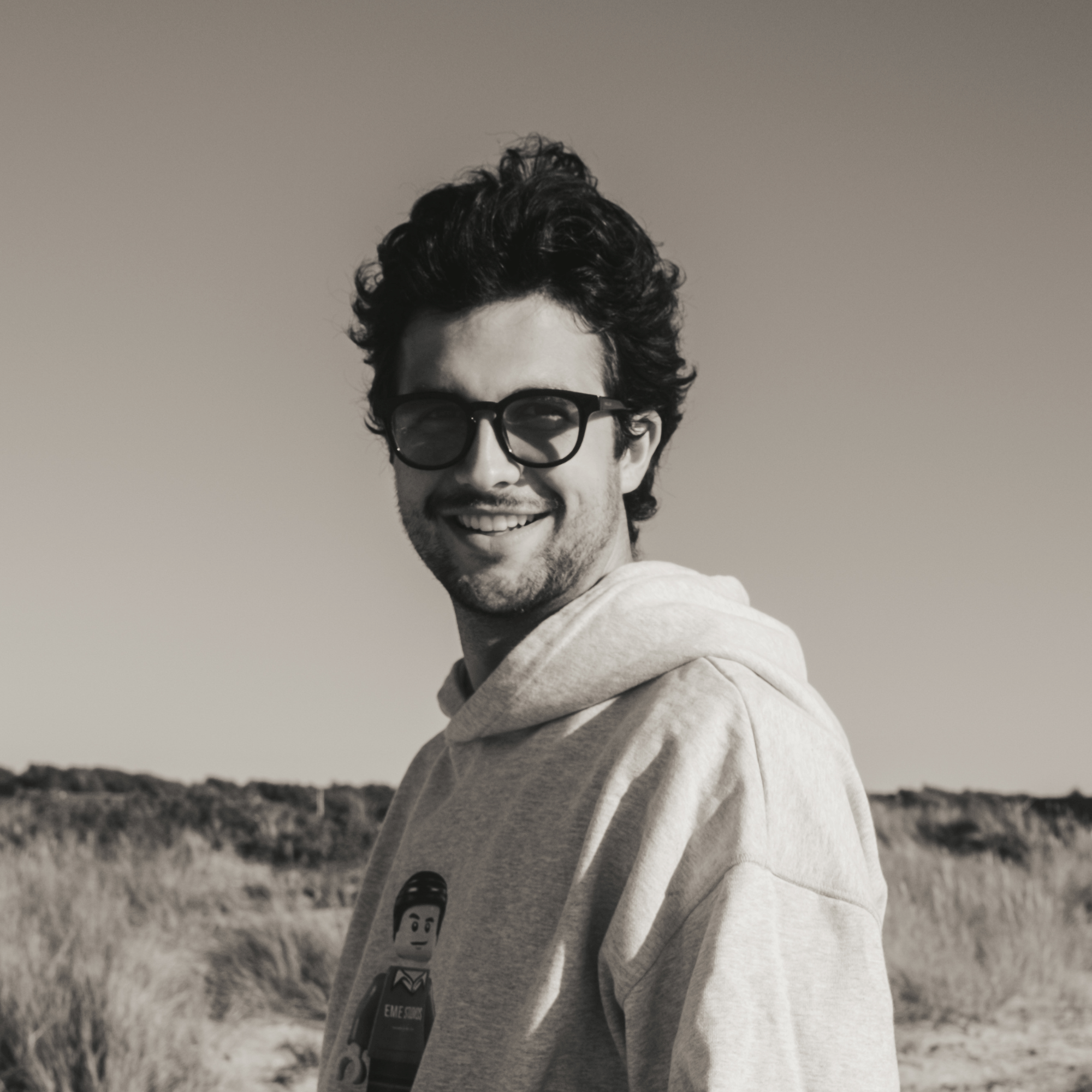 Junger Mann mit dunklen Locken und Brille lächelt, trägt grauen Kapuzenpullover mit Lego-Figur-Design, steht draußen bei sandigem Gelände mit Gras unter klarem Himmel.