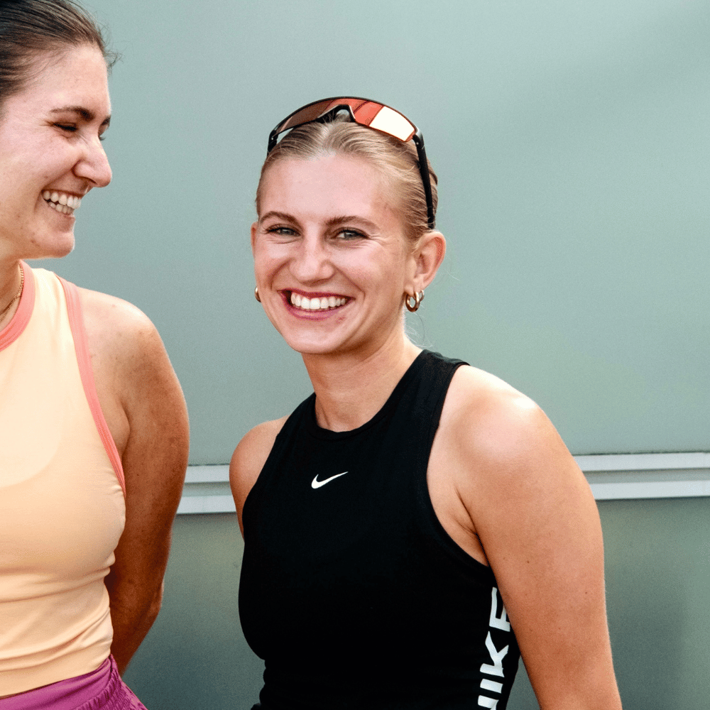 Zwei junge Frauen lächeln in Sportbekleidung, eine trägt ein schwarzes Nike-Top und Sonnenbrille auf dem Kopf, die andere ein helles Top.
