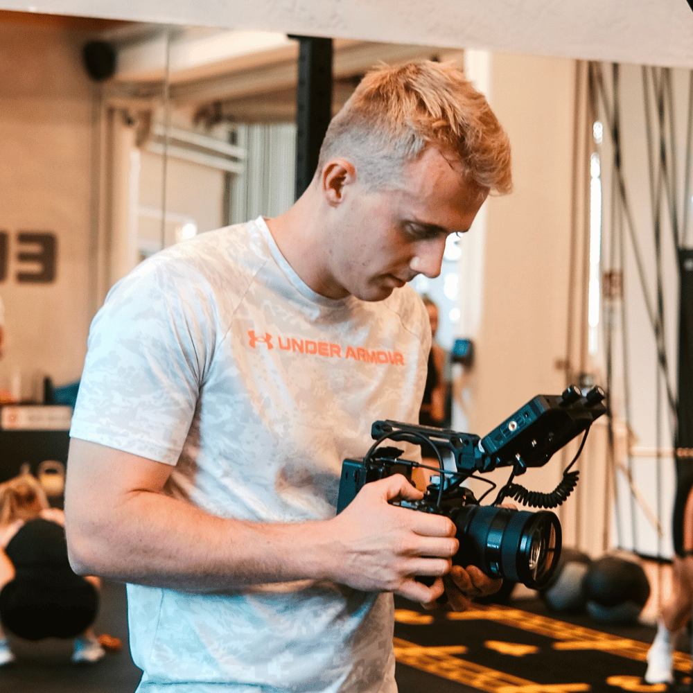 Junger Mann in weißem Under Armour Shirt hält und bedient professionelle Kamera in einem Fitnessstudio.