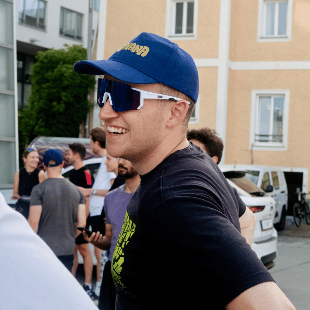 Junger Mann mit blauem Cap und großen, weißen und blauen Sonnenbrillen lacht im Freien, im Hintergrund stehen mehrere stehende Personen vor einem gelblichen Wohngebäude.