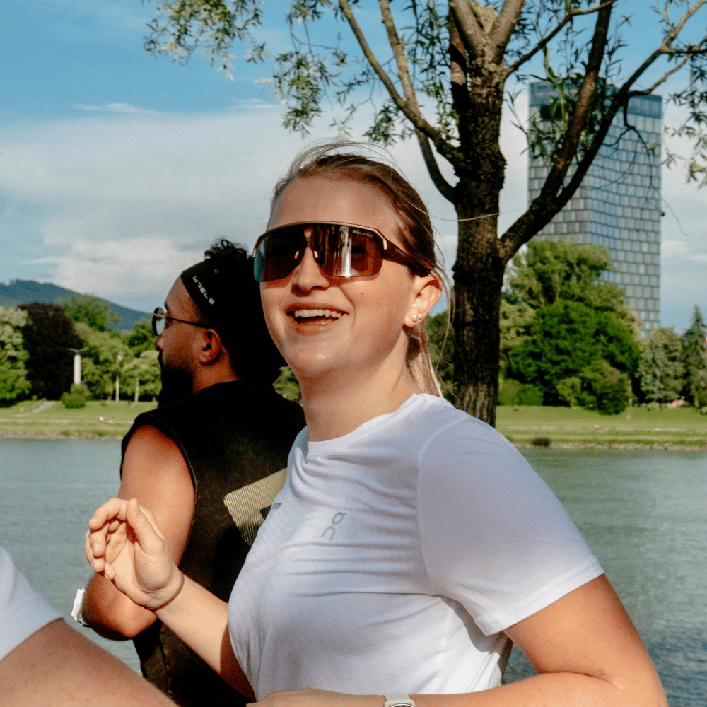 Frau in Sportkleidung und Sonnenbrille lacht beim Joggen neben einem Fluss mit grünen Bäumen und Hochhaus im Hintergrund.