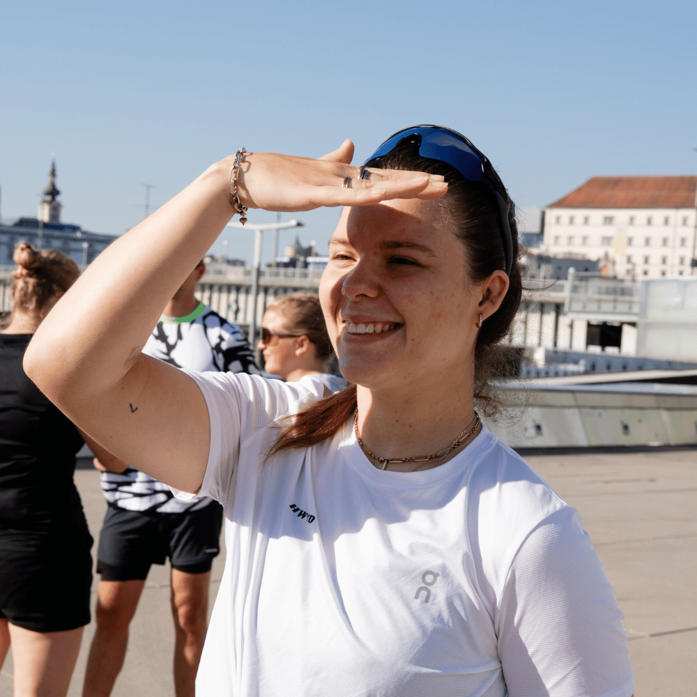 Junge Frau in weißem Sportshirt hält die Hand über die Augen, um gegen die Sonne zu schauen, mit anderen sportlich gekleideten Personen im Hintergrund.