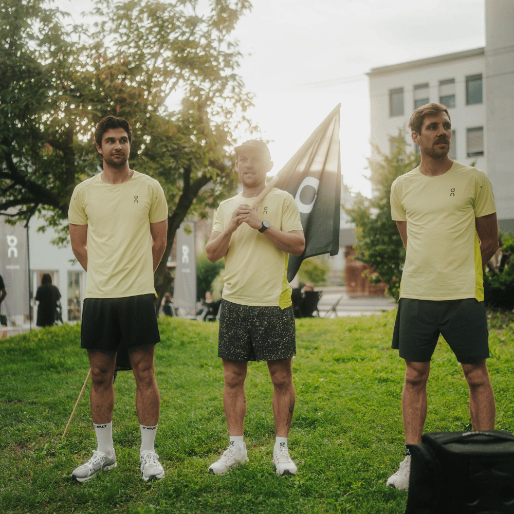 Drei Männer in gelben Sportshirts und Shorts stehen draußen auf grünem Gras. Der Mann in der Mitte hält eine Flagge mit einem Logo, Bäume und Gebäude sind im Hintergrund.
