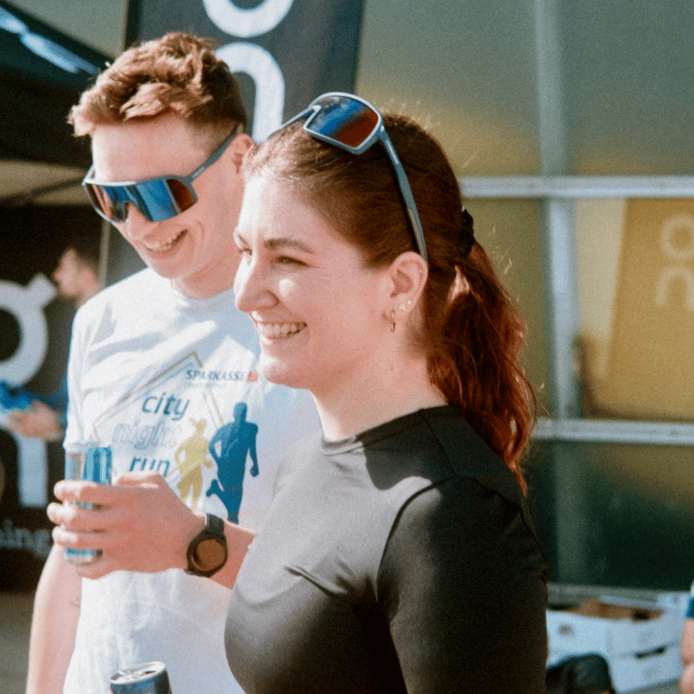 Zwei Personen lachen, beide tragen Sportbrillen. Die Frau hat rote Haare und ein schwarzes Shirt, der Mann trägt ein weißes T-Shirt mit einem City Night Run-Logo und hält eine Dose.