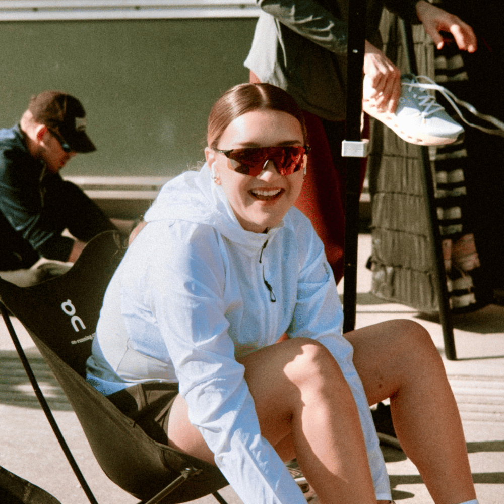 Frau mit Sportbrille und weißer Jacke sitzt lachend auf einem Klappstuhl im Freien, während im Hintergrund eine Person Sportschuhe hält.