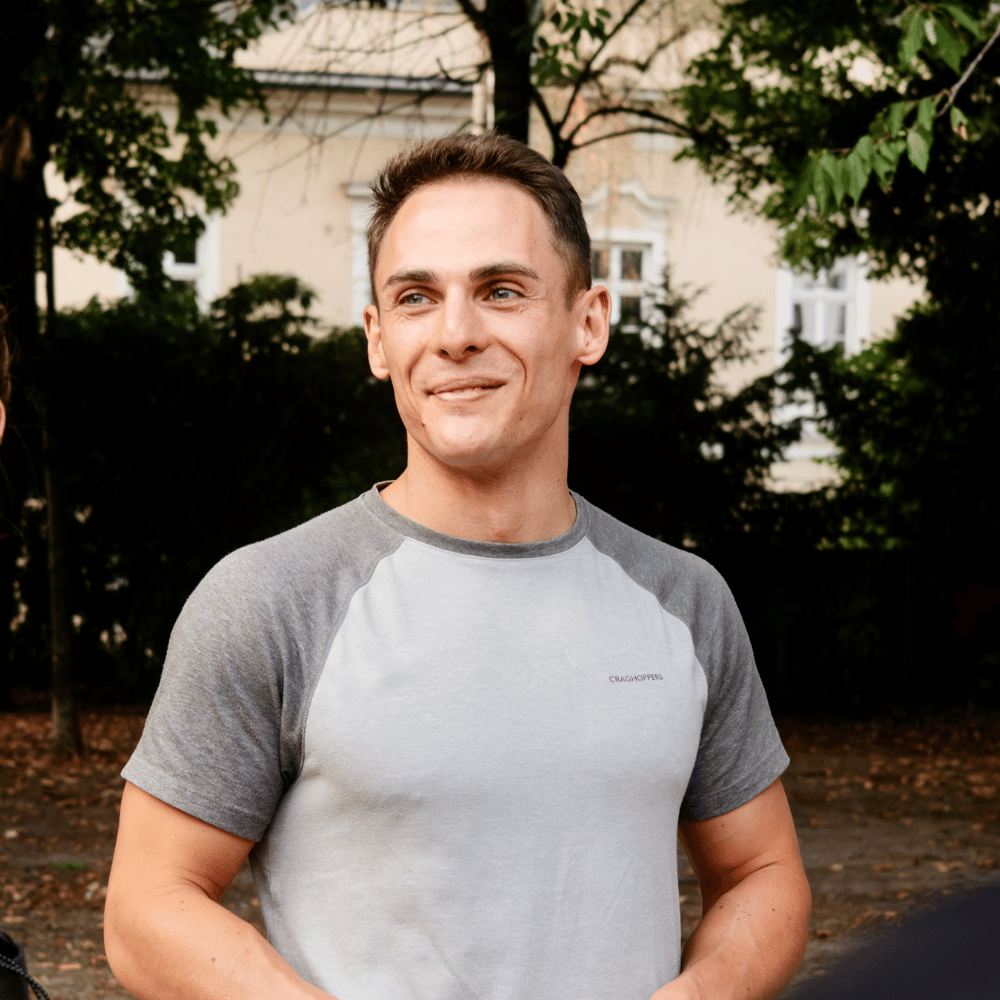 Junger Mann mit grauem T-Shirt steht draußen vor Bäumen und einem hellen Gebäude, er lächelt und schaut seitlich.