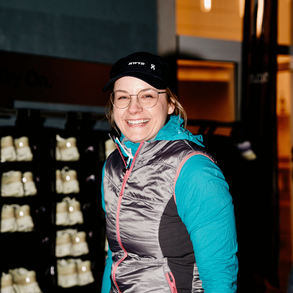 Lächelnde Frau mit Brille, schwarzer Kappe und silberner Weste über türkisfarbenem Langarmshirt vor Regalen mit mehreren weißen Skischuhen.