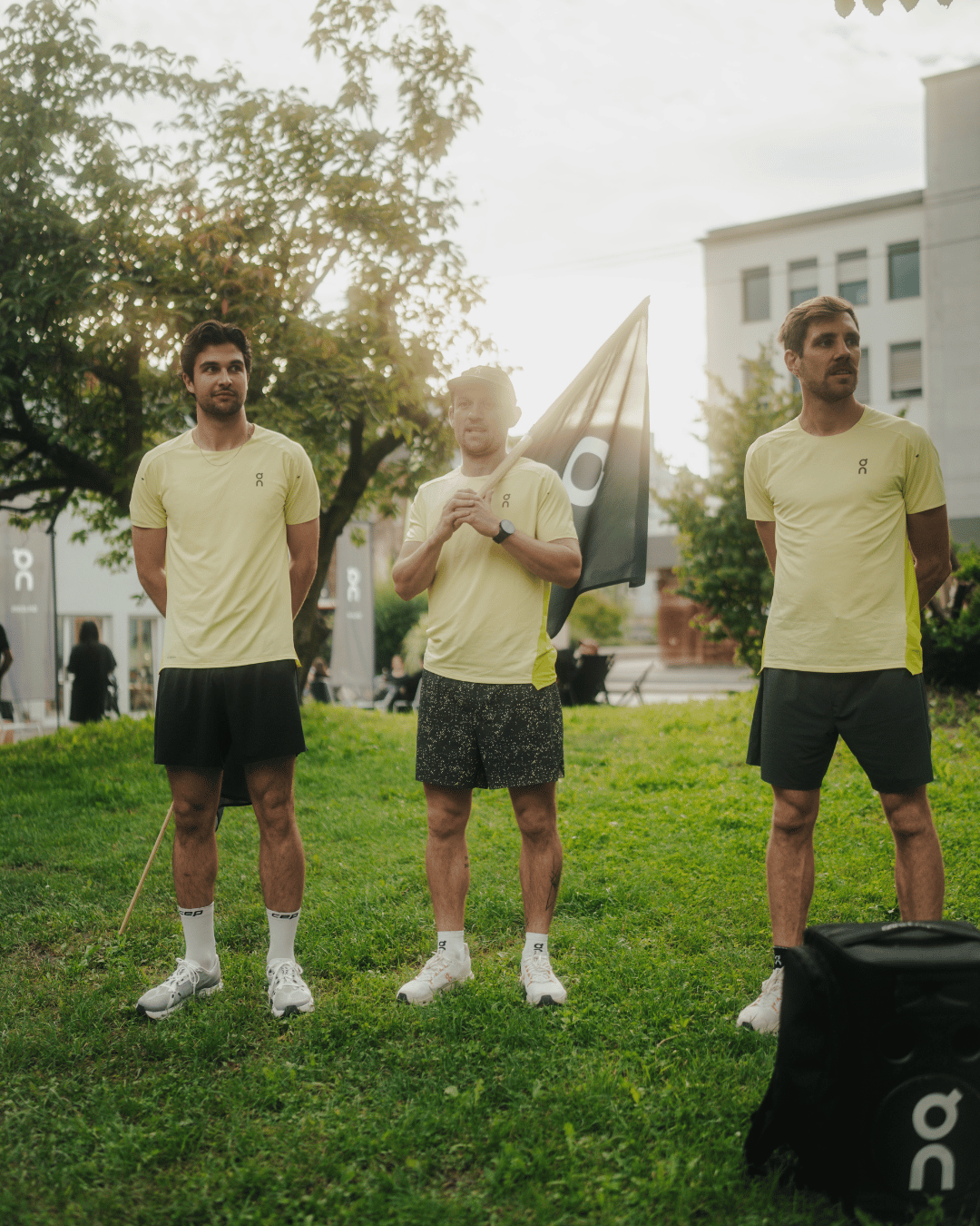 Drei Männer in gelben Sportshirts und schwarzen Shorts stehen auf einer Wiese, einer hält eine Flagge, im Hintergrund Bäume und Gebäude bei Sonnenlicht.