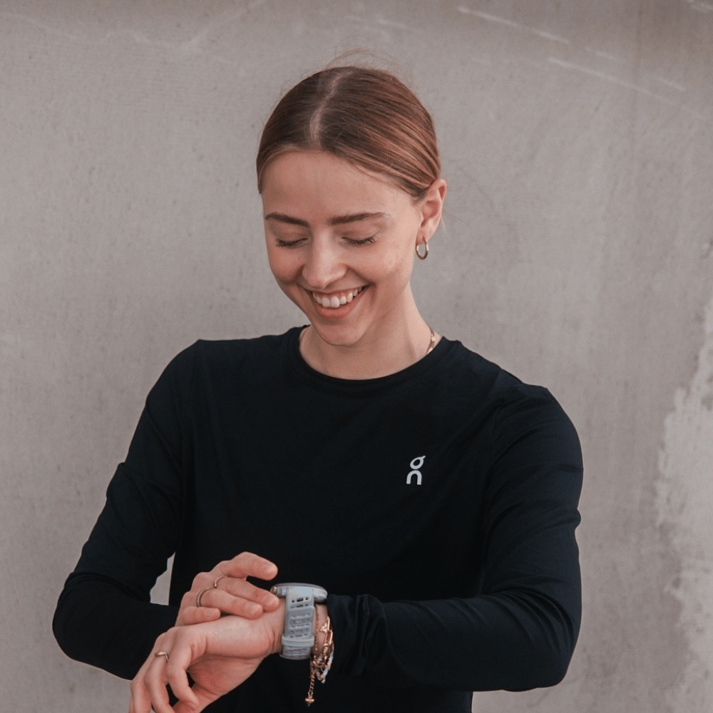 Eine Frau mit einem schwarzen Langarmshirt sieht lächelnd auf eine Smartwatch an ihrem linken Handgelenk, während sie vor einer grauen Wand steht.