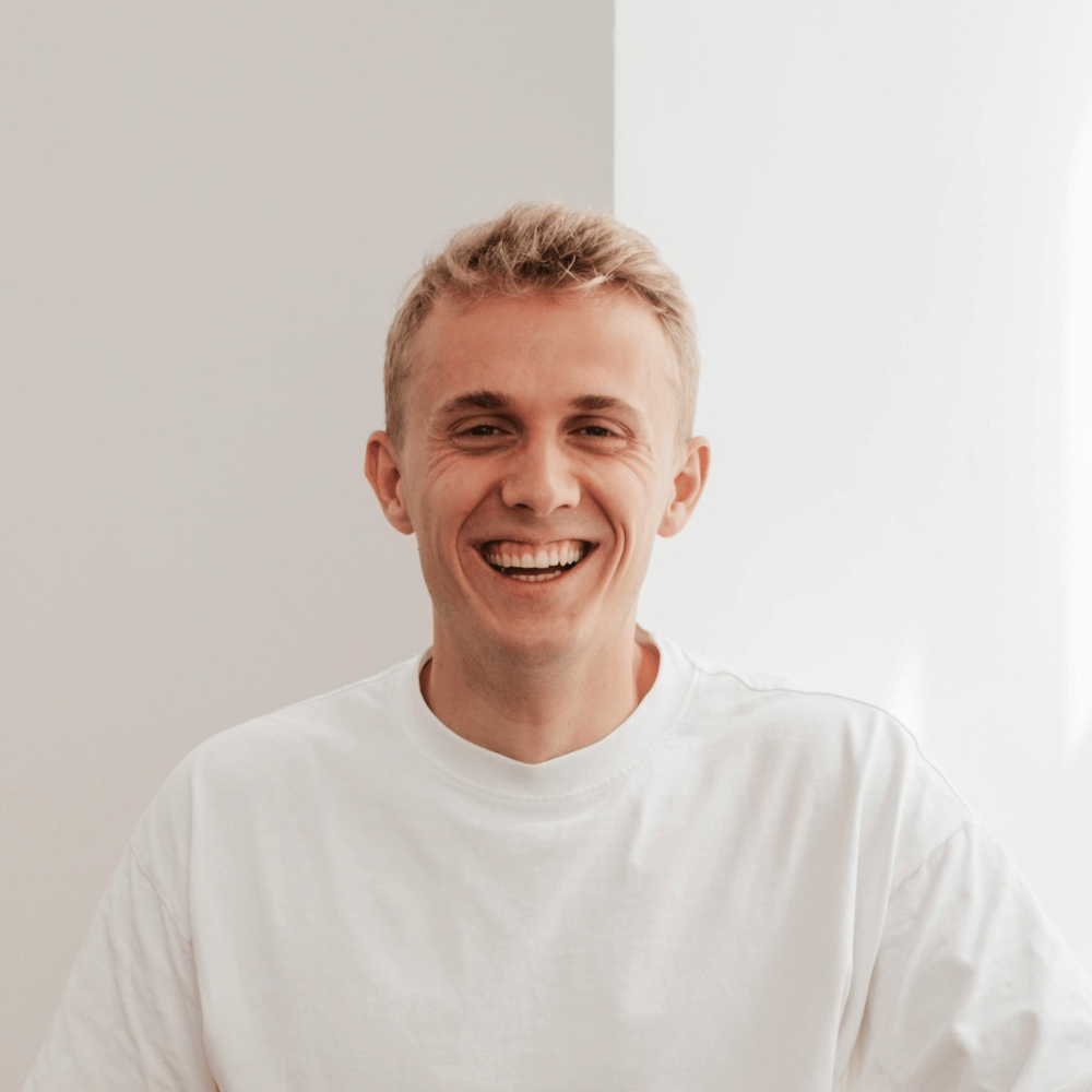 Junger Mann mit kurzen blonden Haaren, der lächelnd einen weißen T-Shirt trägt vor einer zweifarbigen Wand.