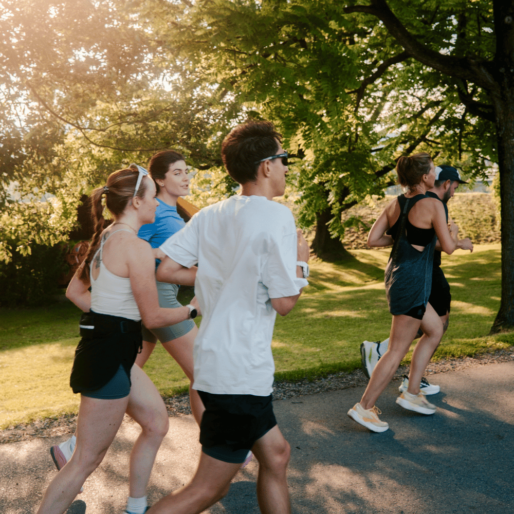 Fünf Menschen joggen zusammen in einem sonnigen Park, umgeben von grünen Bäumen und Gras, sie tragen Sportkleidung und genießen die Bewegung im Freien.