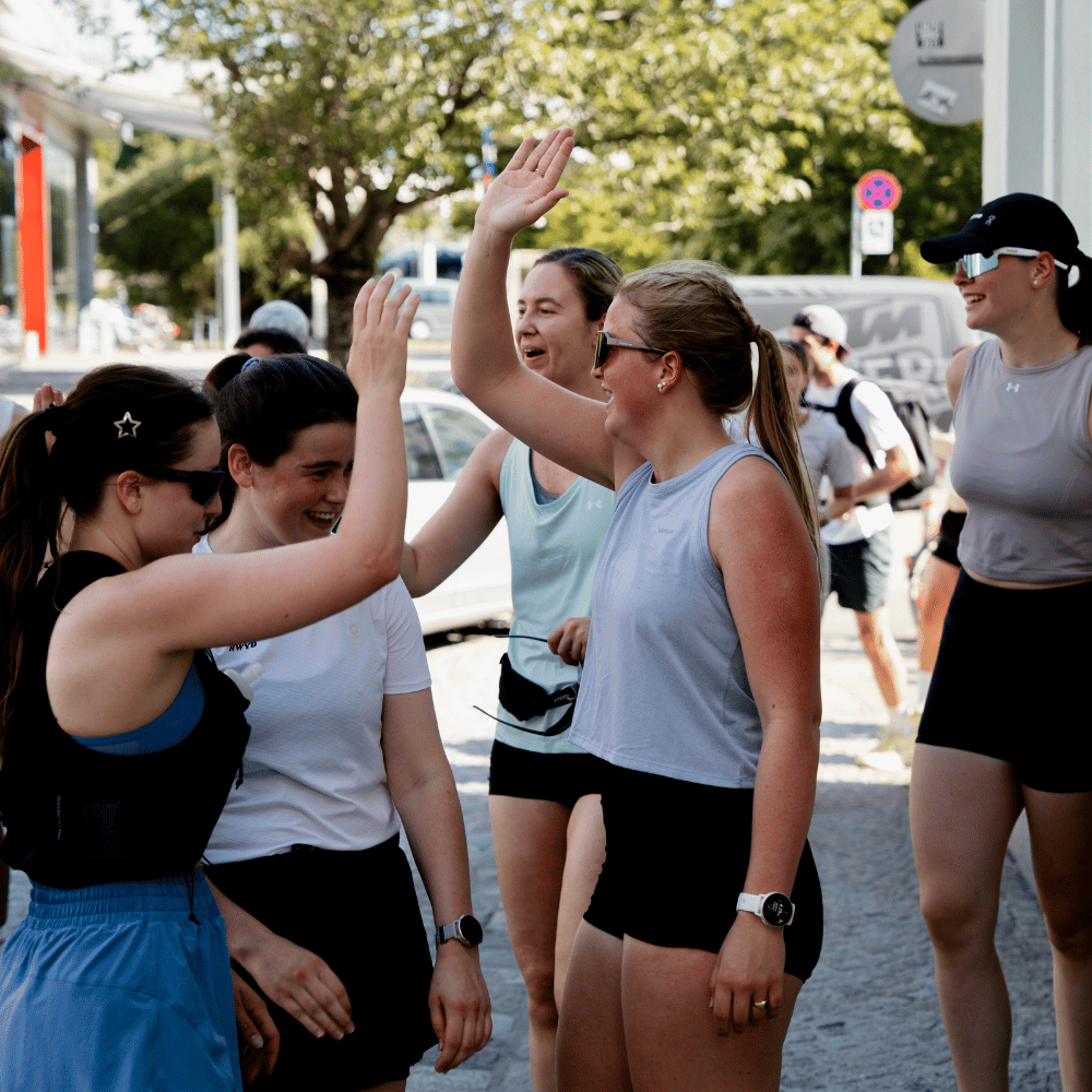 Gruppe junger Frauen in Sportbekleidung, die draußen stehen und sich zur Begrüßung die Hände zum High-Five heben, umgeben von Bäumen und parkenden Autos.