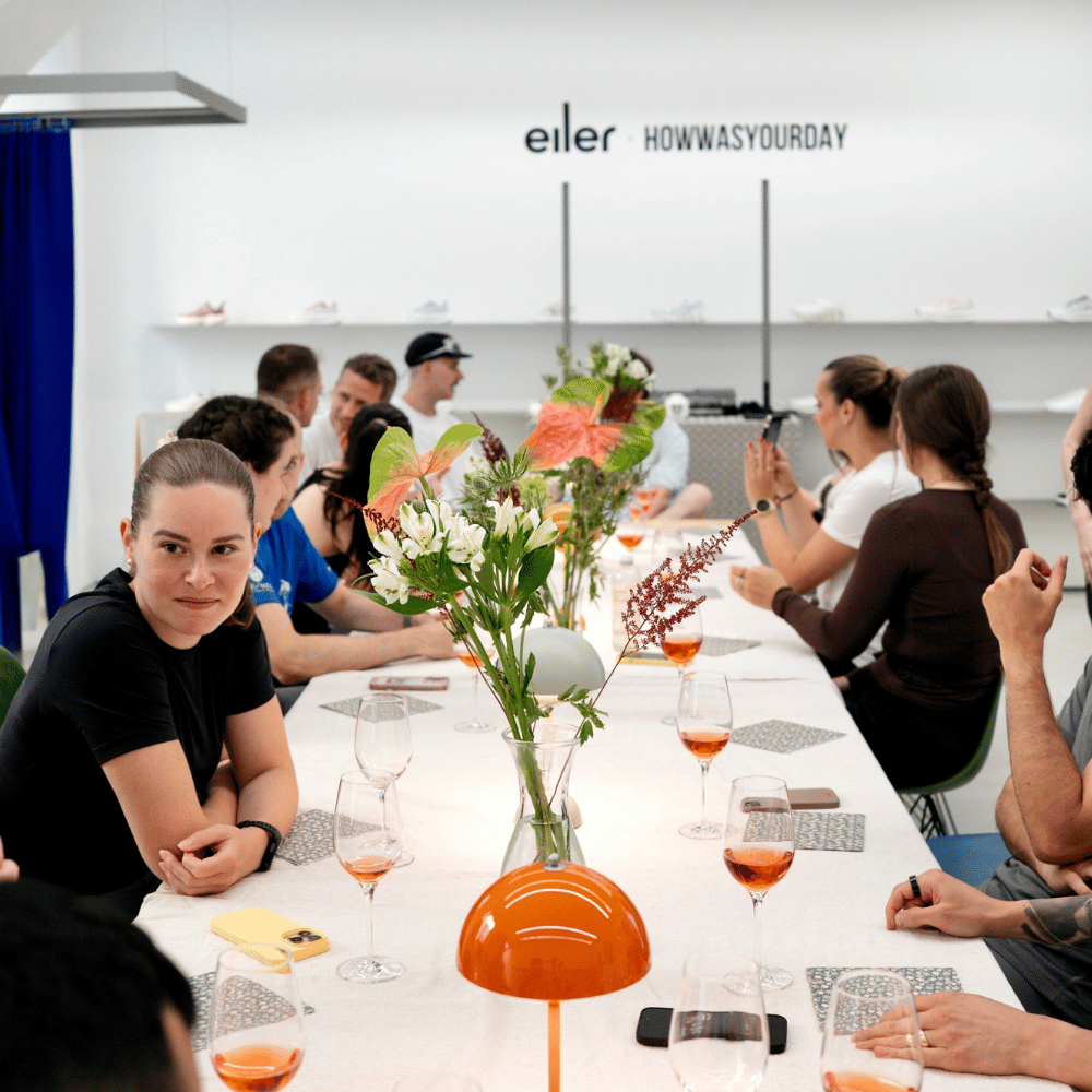 Menschen sitzen um einen langen Tisch mit Blumen und Gläsern mit orangefarbenem Getränk, im Hintergrund Logo mit Text 'eller HOWWASYOURDAY' an weißer Wand.