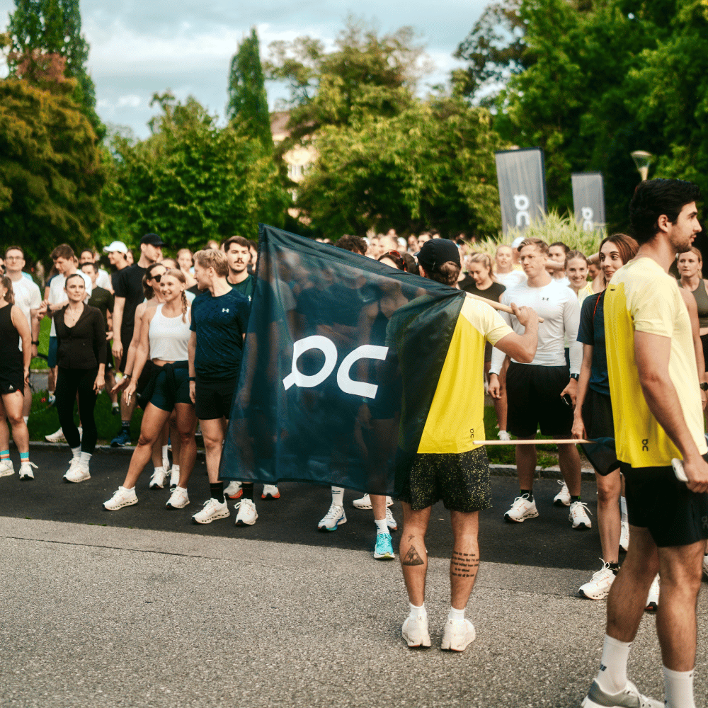 Eine Gruppe Läufer versammelt auf einer Straße im Freien, zwei Männer in gelben Shirts halten eine schwarze Fahne mit weißem 'on'-Logo, Bäume sind im Hintergrund sichtbar.