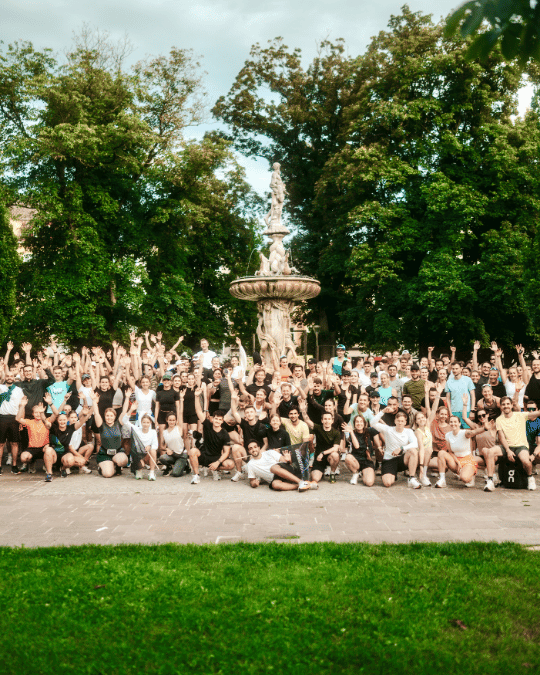 Große Gruppe von Menschen posiert vor einem dekorativen Springbrunnen in einem Park, viele heben die Arme, um den Moment zu feiern.