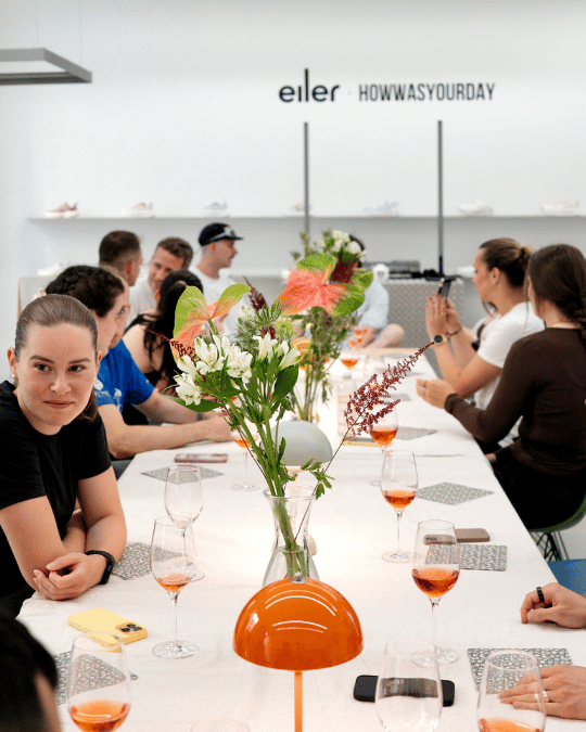 Menschen sitzen an einem langen Tisch mit Blumen in der Mitte und Gläsern mit roséfarbenem Getränk, in einem hellen Raum mit der Aufschrift „eiler HOWWASYOURDAY“ an der Wand.