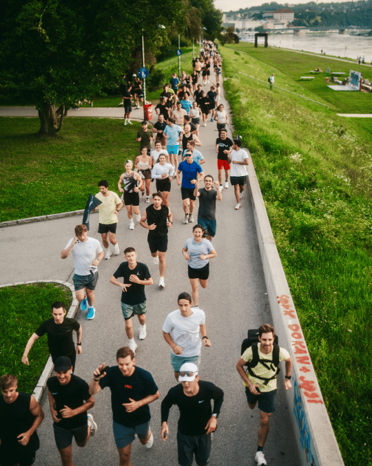 Gruppe von Menschen verschiedenen Alters läuft auf einem Gehweg neben einem Fluss mit grünem Gras und Bäumen im Hintergrund.