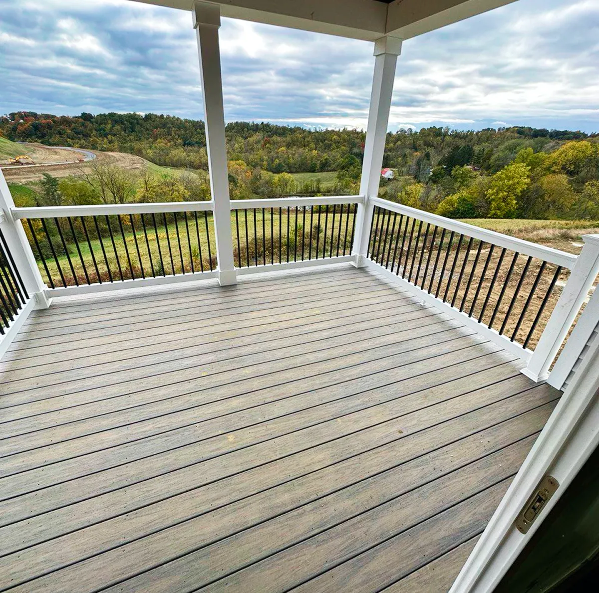 View from an empty covered wooden deck with white posts and black railings overlooking a grassy landscape with trees under a cloudy sky.