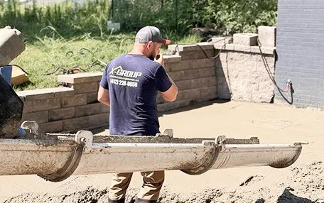 Construction worker wearing a blue shirt and gray cap stands near a concrete pouring chute as fresh concrete is spread on the ground beside a brick retaining wall.