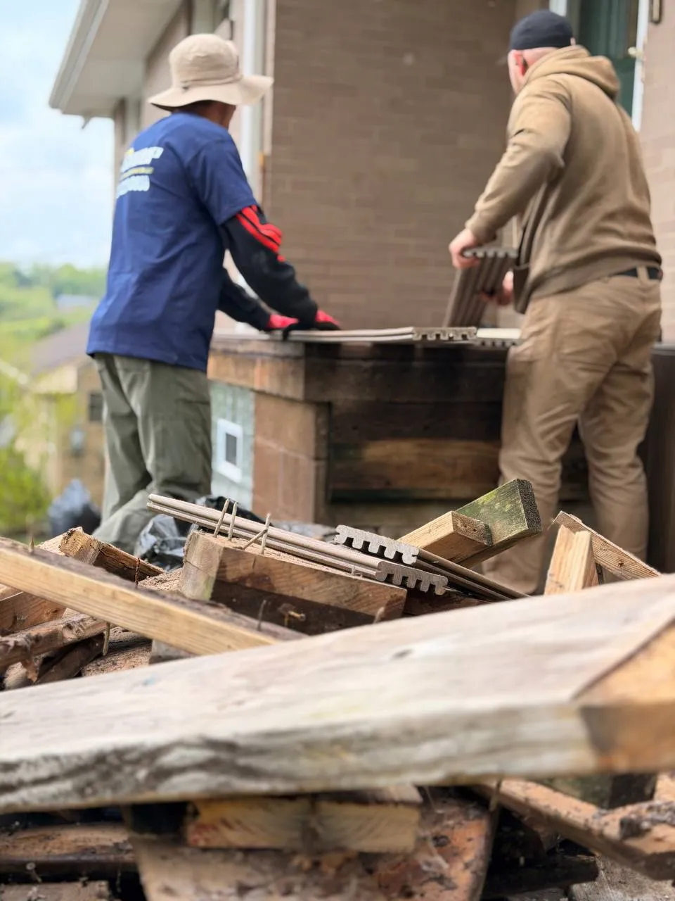 Two workers handling deck materials near a brick house, with a pile of wooden debris and deck boards in the foreground.