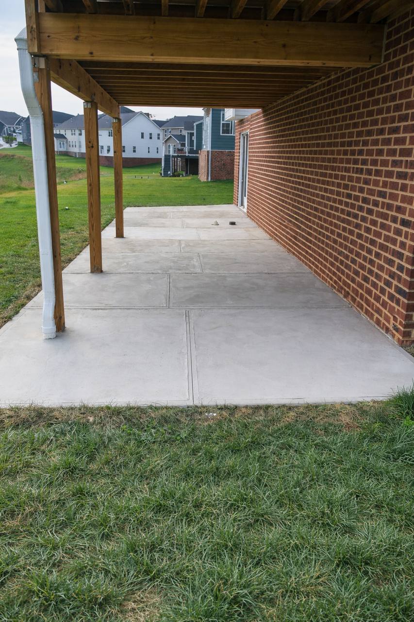 Concrete patio underneath a wooden deck attached to a brick house with green grass lawn surrounding the patio.