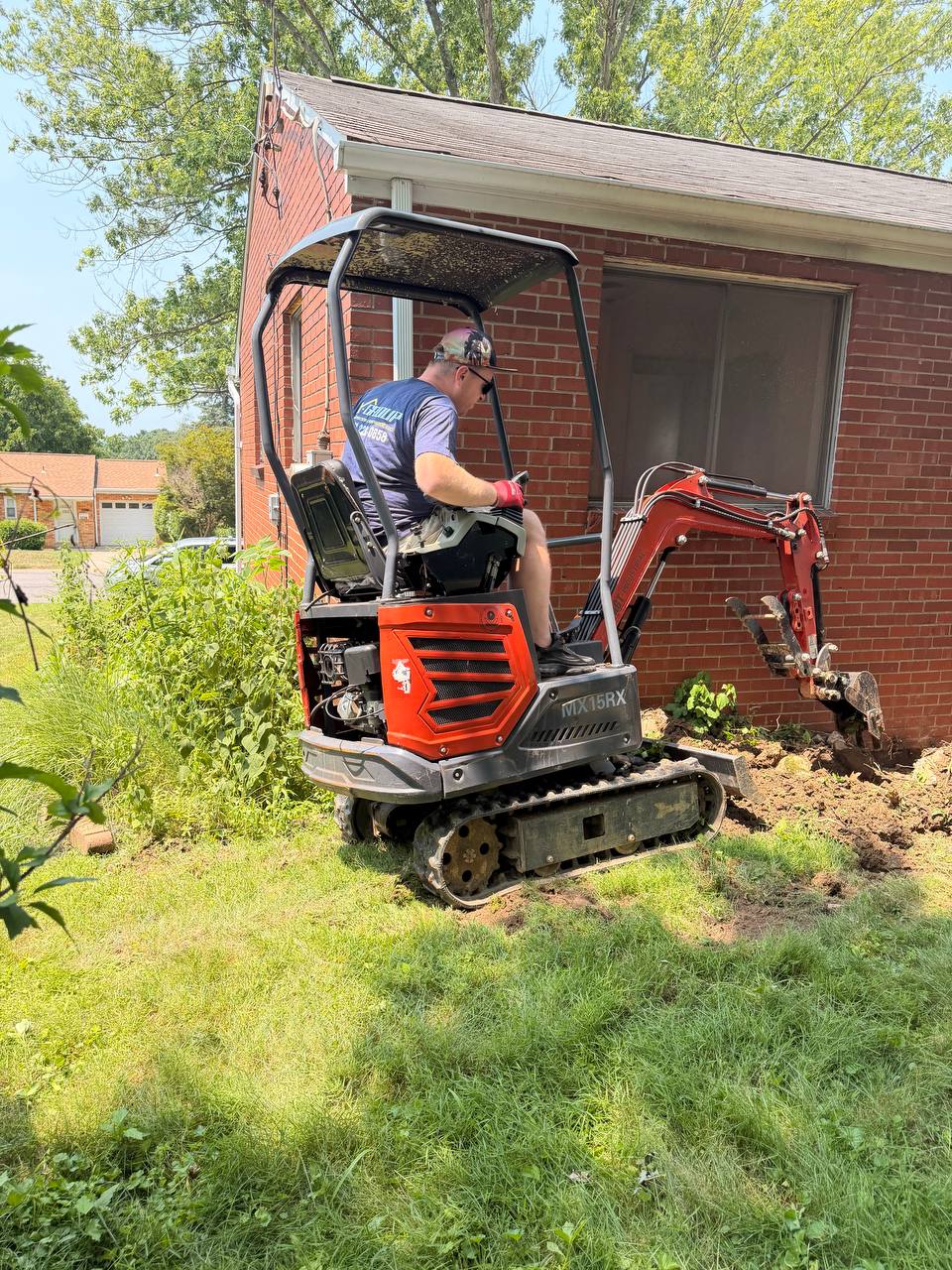 Man operating a small orange and black excavator with tracks, digging soil next to a red brick house with a window, on a grassy lawn with green plants nearby.