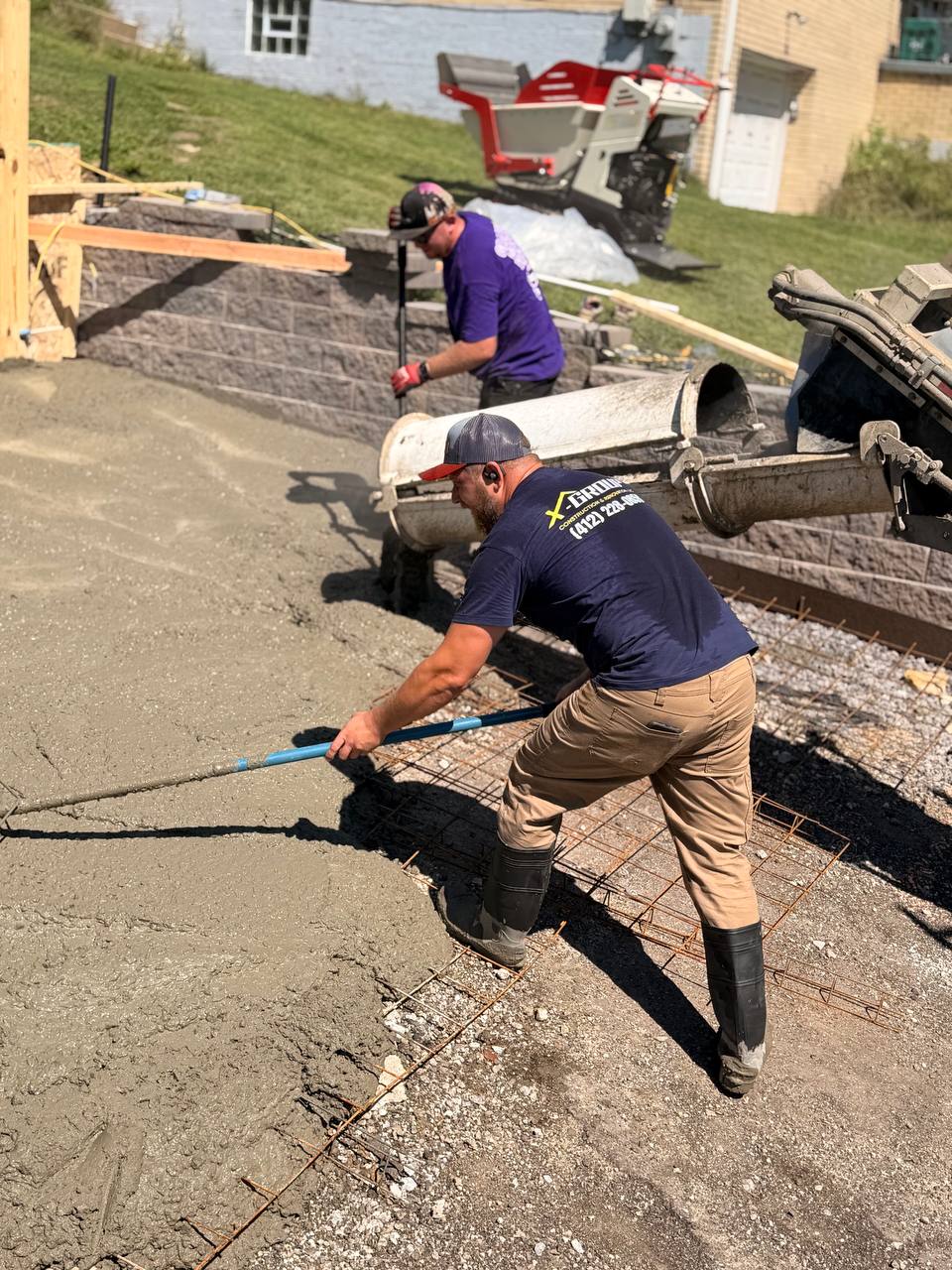 Two construction workers pouring and spreading concrete over metal rebar on a driveway near a house, with one worker smoothing the wet concrete using a tool.