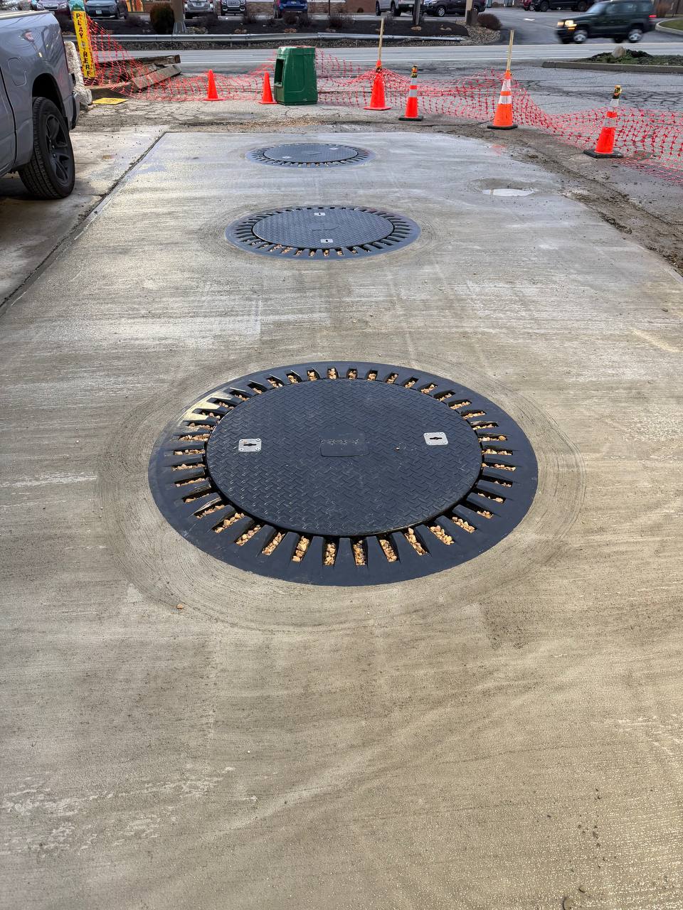 Concrete surface with three round black metal utility covers embedded in a row; gravel fills slots around each cover. Orange safety cones and red mesh fencing separate the area from the street in the background, with parked cars visible beyond.