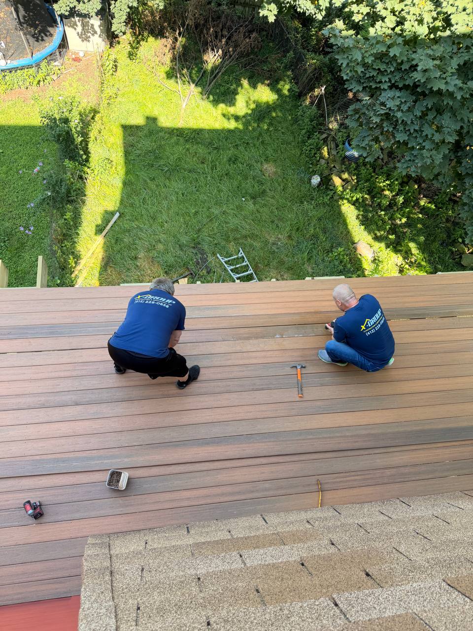 Two men wearing blue shirts are working on building or repairing a wooden elevated deck, viewed from above, with tools like a hammer and a drill beside them and a grassy backyard below.